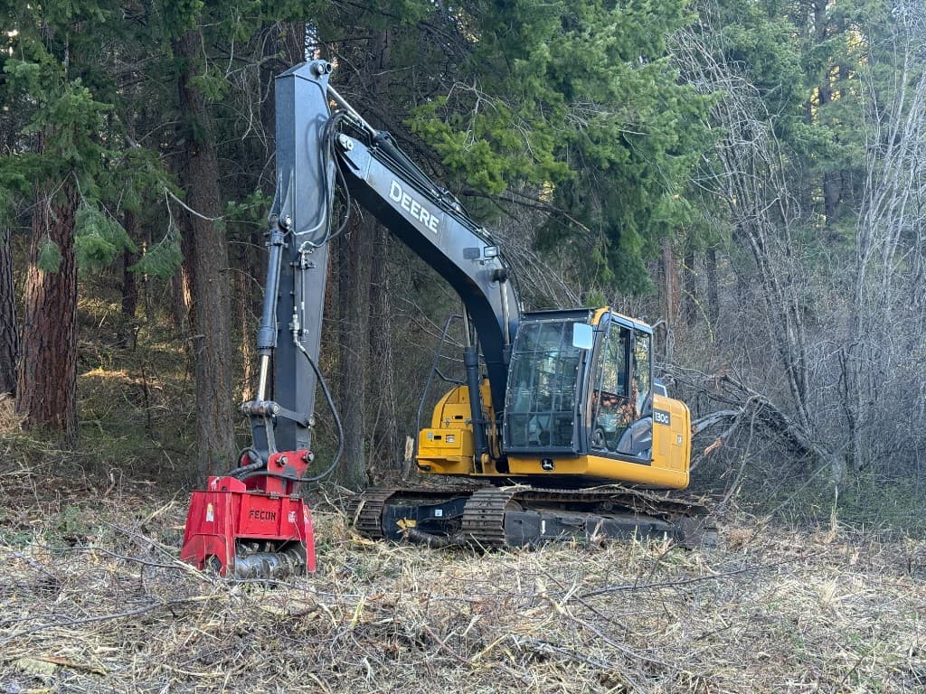 John Deere excavator with Fecon mulching head clearing brush and small timber at a forest edge